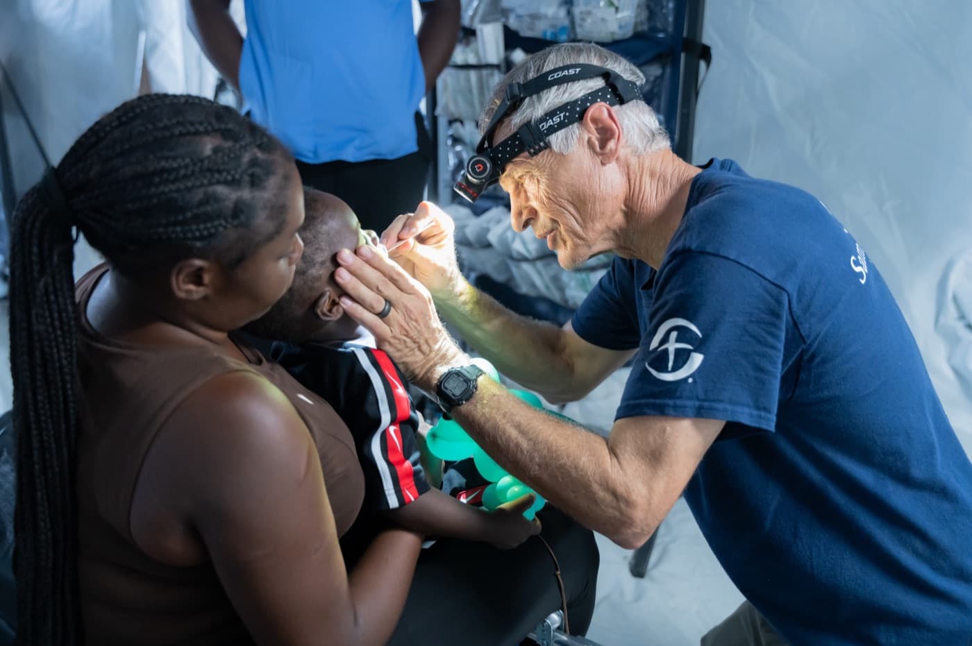 A doctor with a headlamp examines a child’s eyes in a treatment tent.